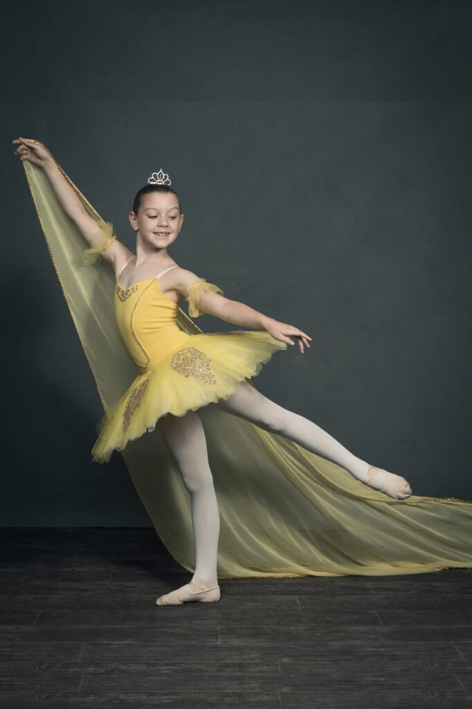 A smiling girl in a ballet pose, wearing a yellow dress and a crown, with a flowing yellow cape.